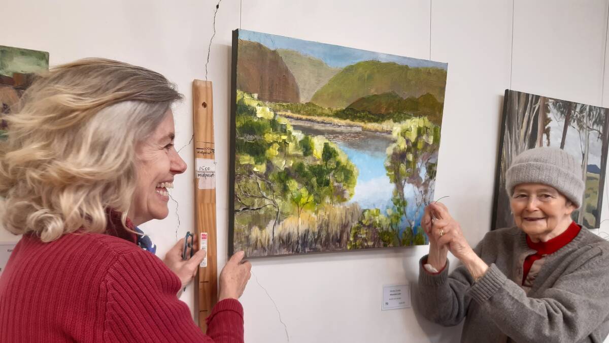 Philippa Charley-Briggs and Judy Wilford hang the artwork: 'Mountain Lake' by Shirley Cooke.